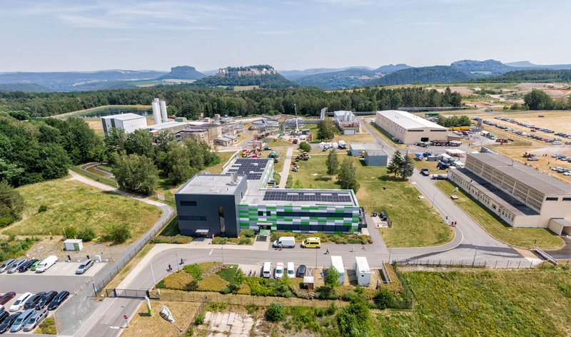 Luftbild mit Gebäuden und technischen Anlagen mit der Festung Königstein und Felsen im Hintergrund