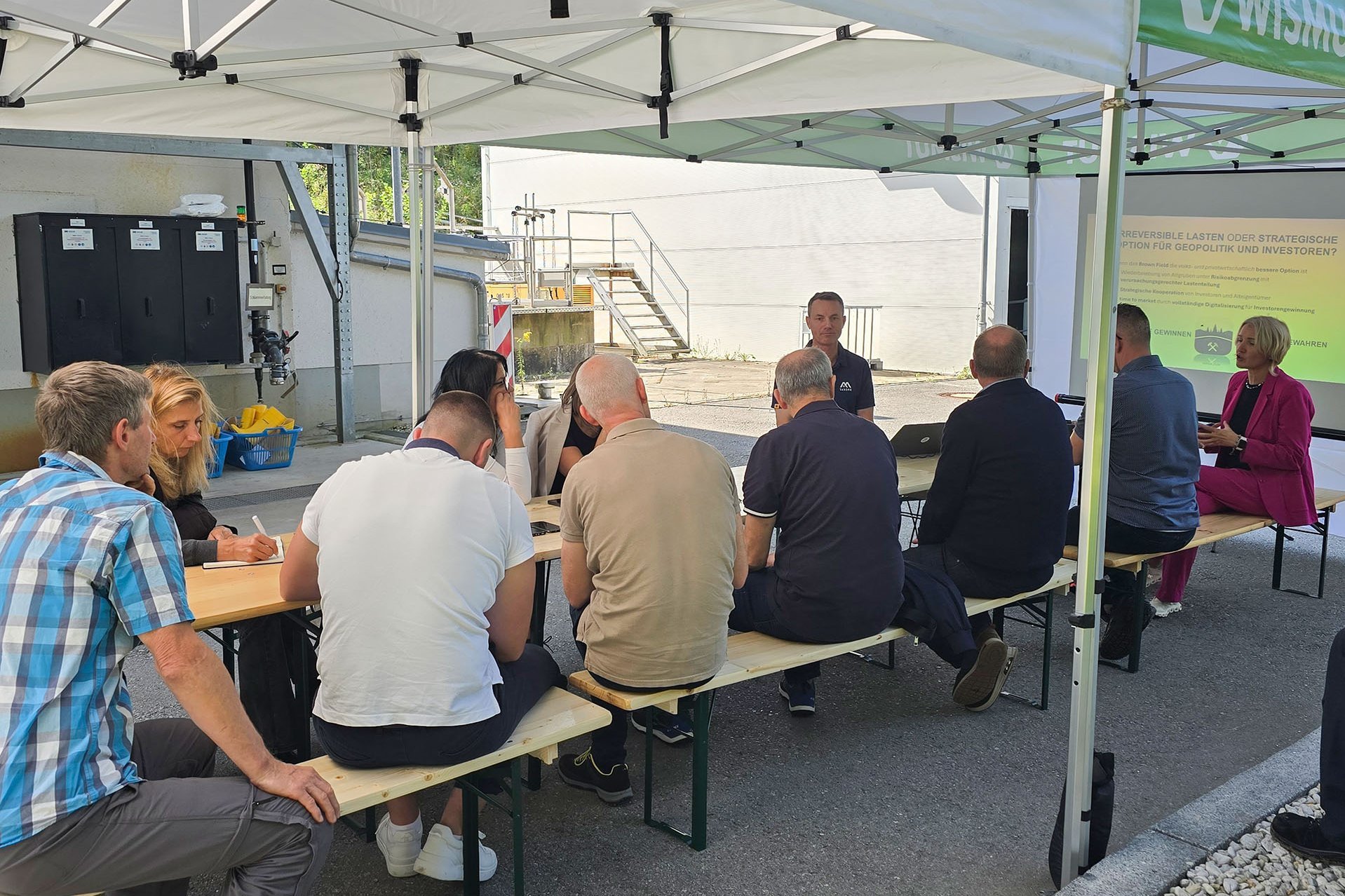 Eine Gruppe sitzt unter einem Pavilion und lauscht einem Bildschirm unterstützten Vortrag