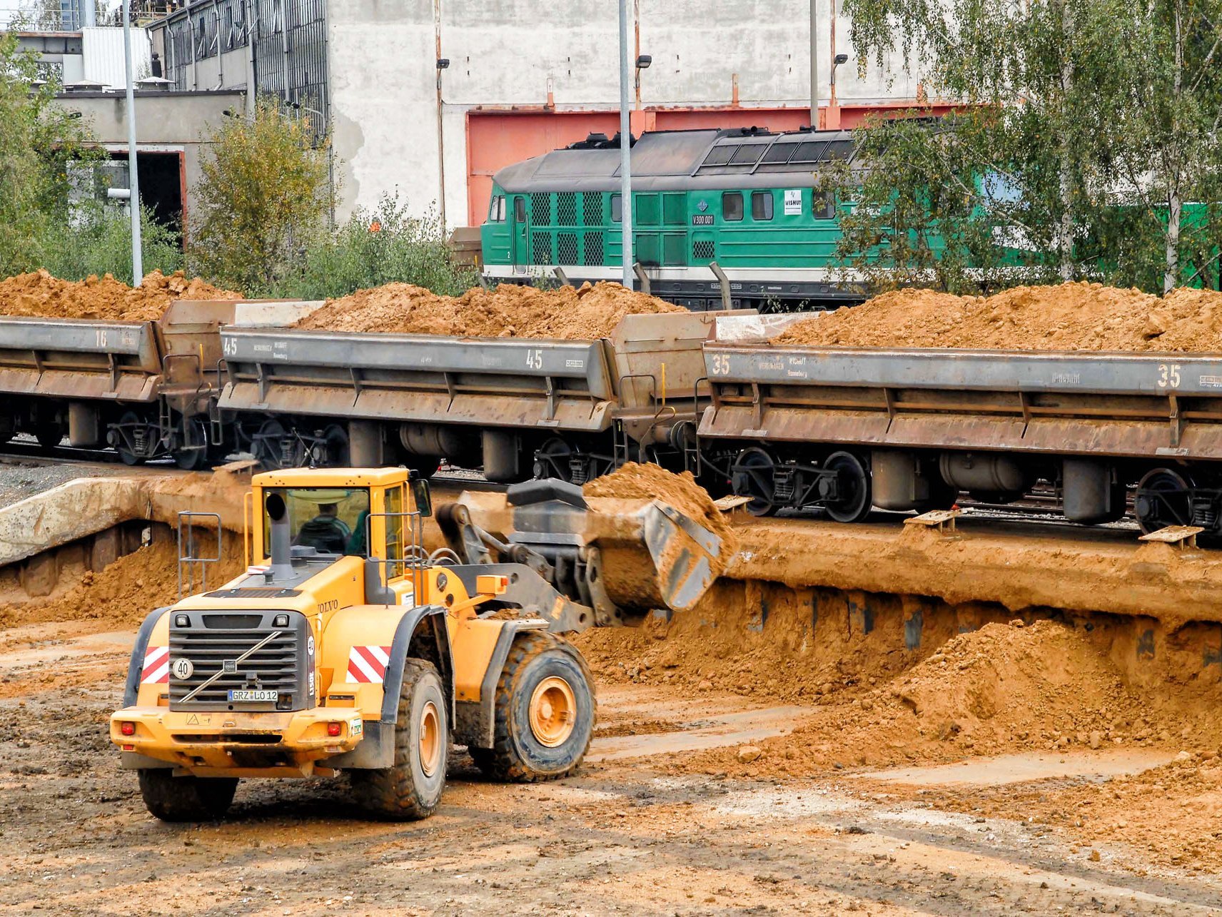 Wagons mit Erdmaterial stehen auf einer Rampe. Ein Radlader nimmt mit der Schaufel abgekipptes Material auf.