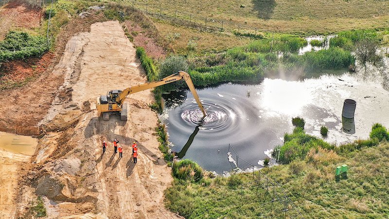 Luftbild: Ein Bagger auf einem Damm greift ins Wasser. Arbeiter scheuen zu.