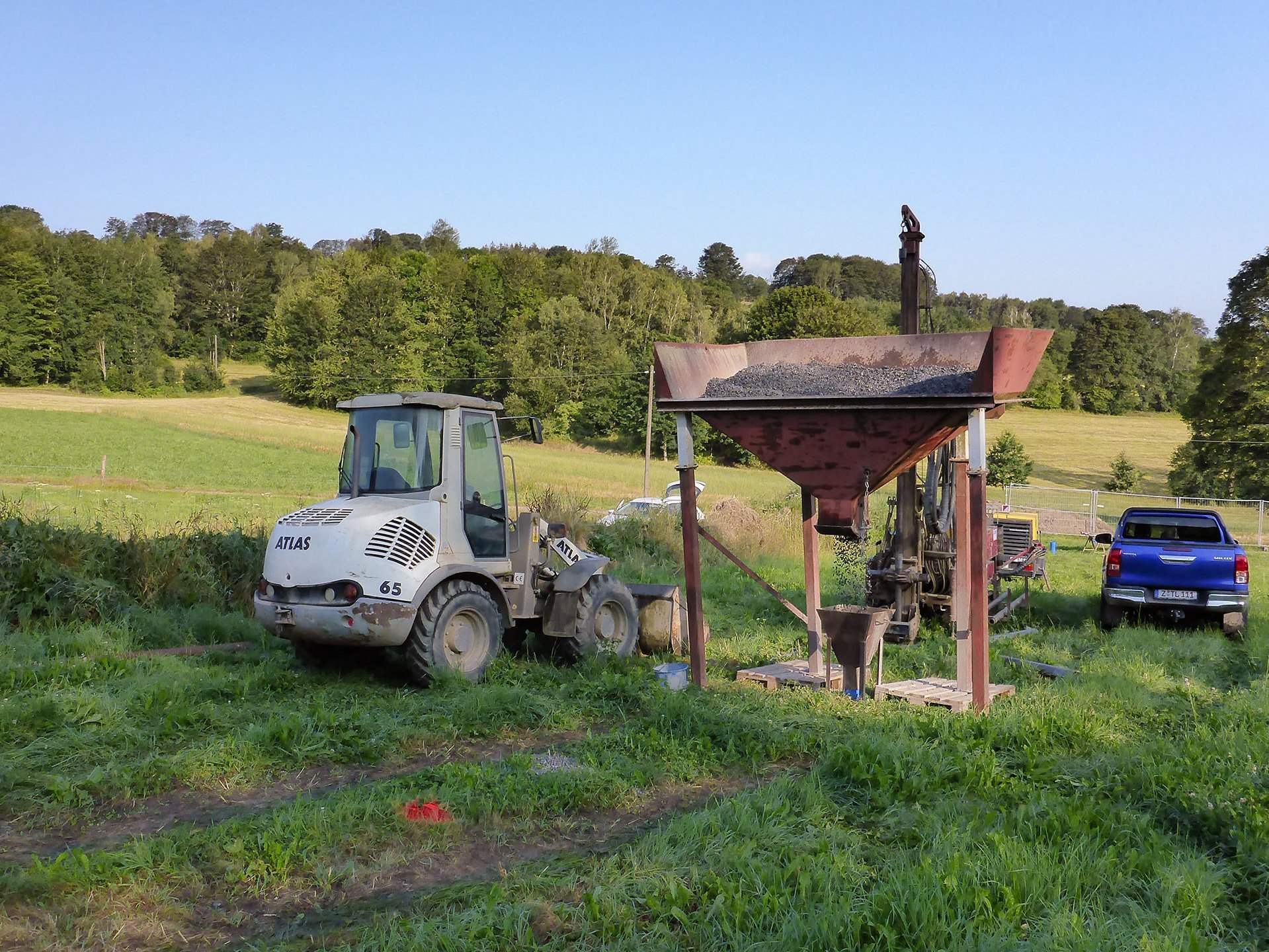 Ein kleiner Radlader steht auf einer Wiese an einem Einfülltrichter an einem Bohrloch
