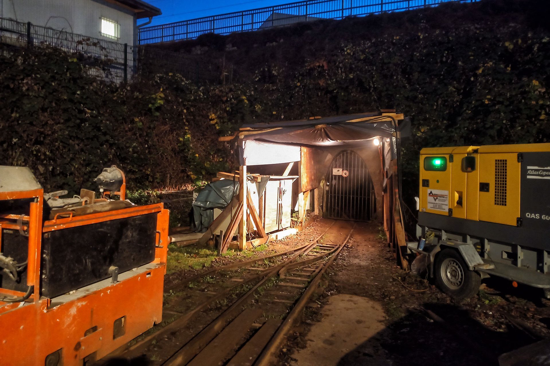 Eine Grubenlok und ein Kompressor vor dem Mundloch in Dämmerlicht