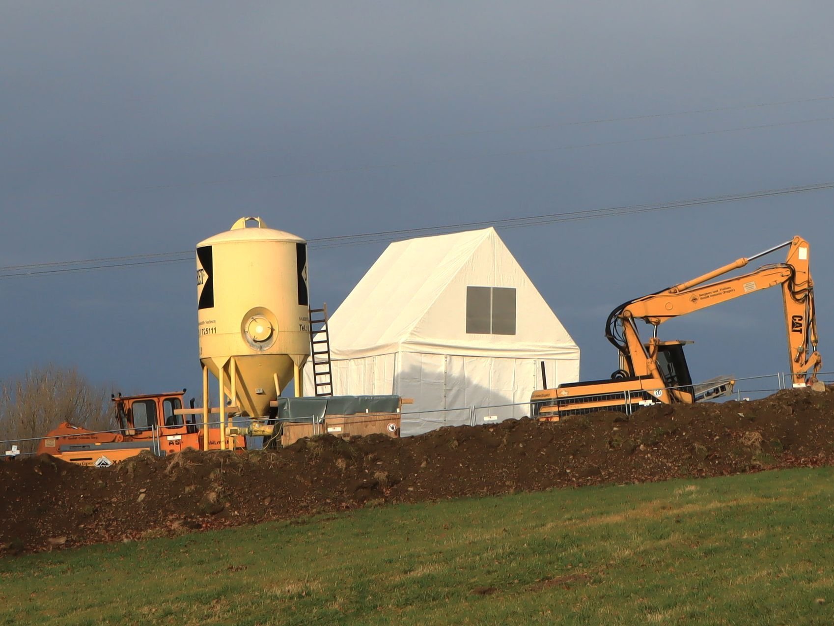 Baustoffsilo, Zelt und Baumaschinen stehen auf einem Feld
