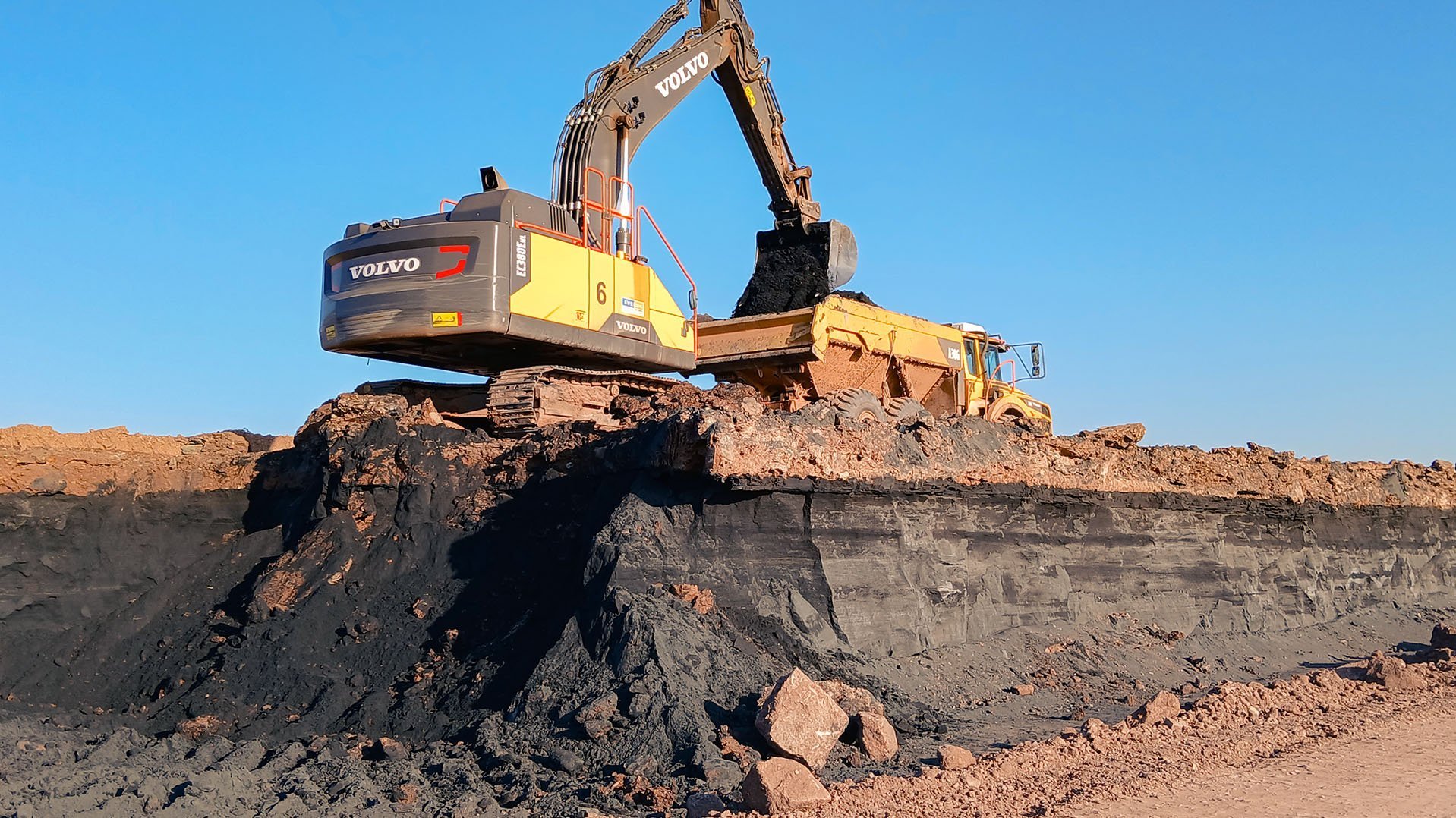 Ein Bagger steht oben an einer Kante schwarzer Tailings und belädt einen Dumper