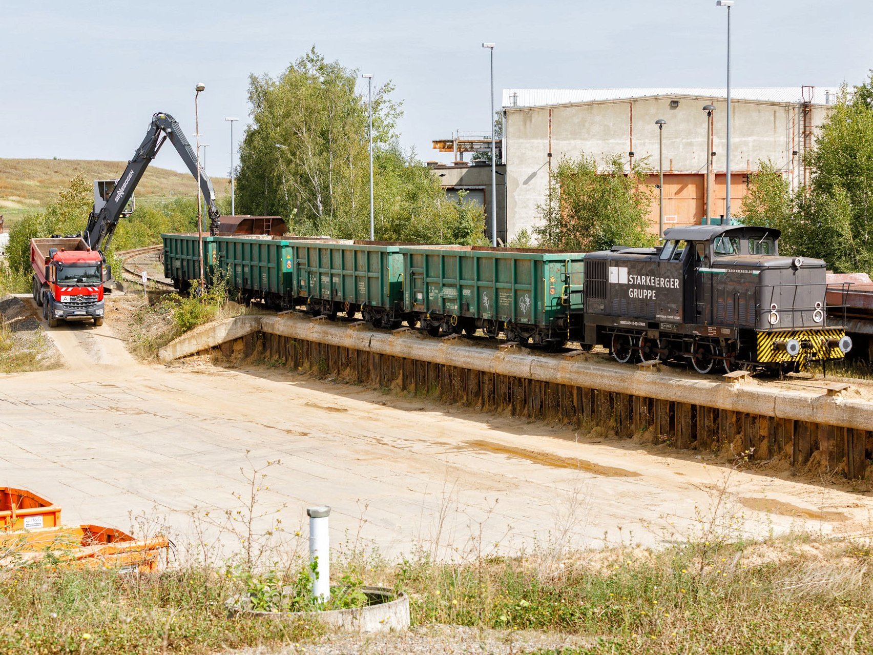 Eine Rangierlok mit drei hohen Wagons steht auf einer Rampe. Ein Bagger entlädt einen Wagon.