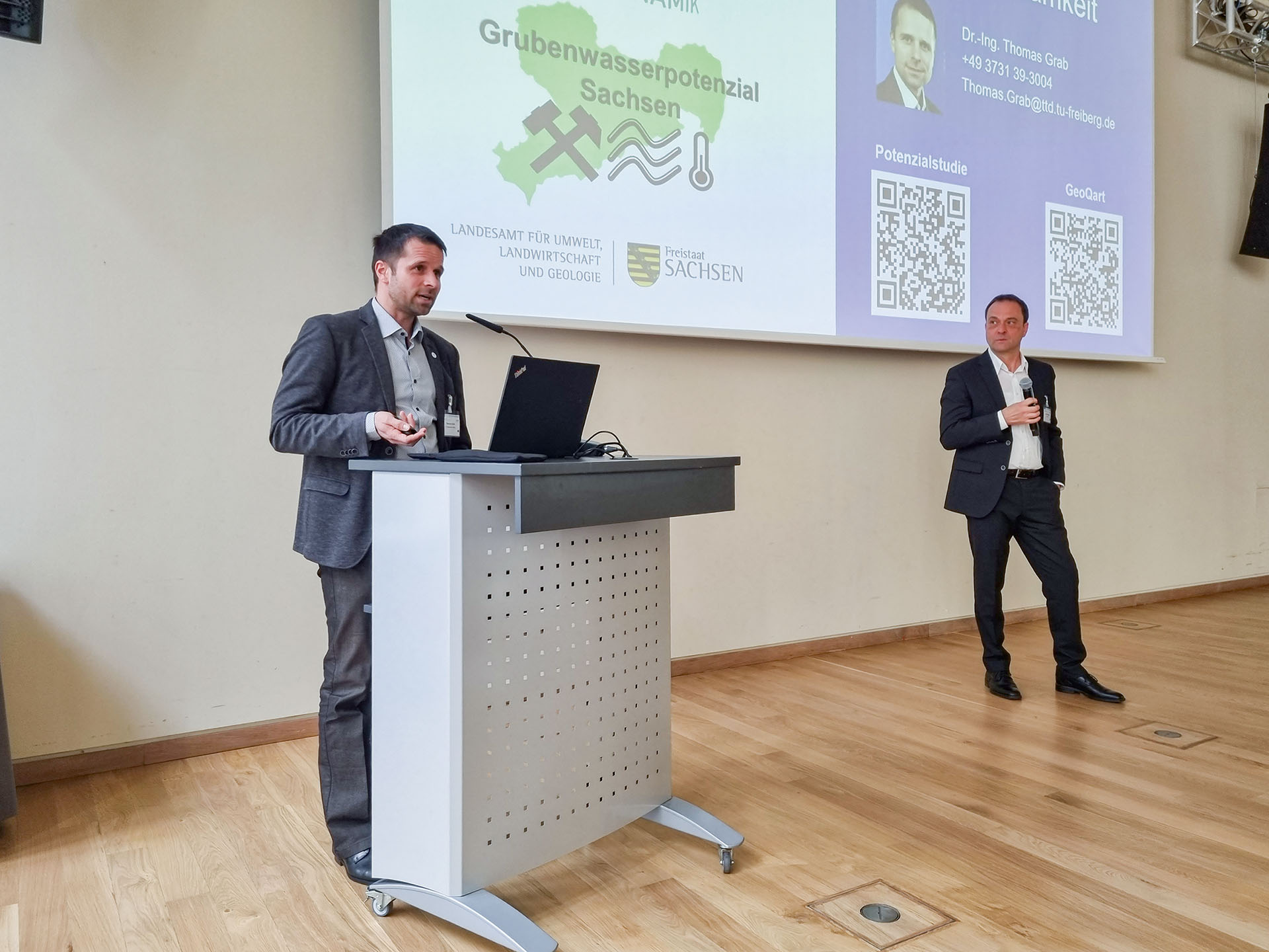 Ein Mann am Rednerpult, ein Mann mit Mikrofon daneben vor einer Präsentation