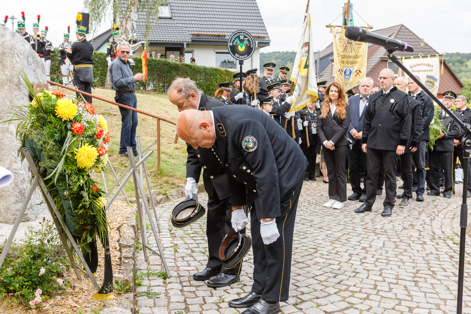 Zwei Männer in Habit ziehen die Mützen und verneigen sich tief vor einem Kranz am Gedenkstein