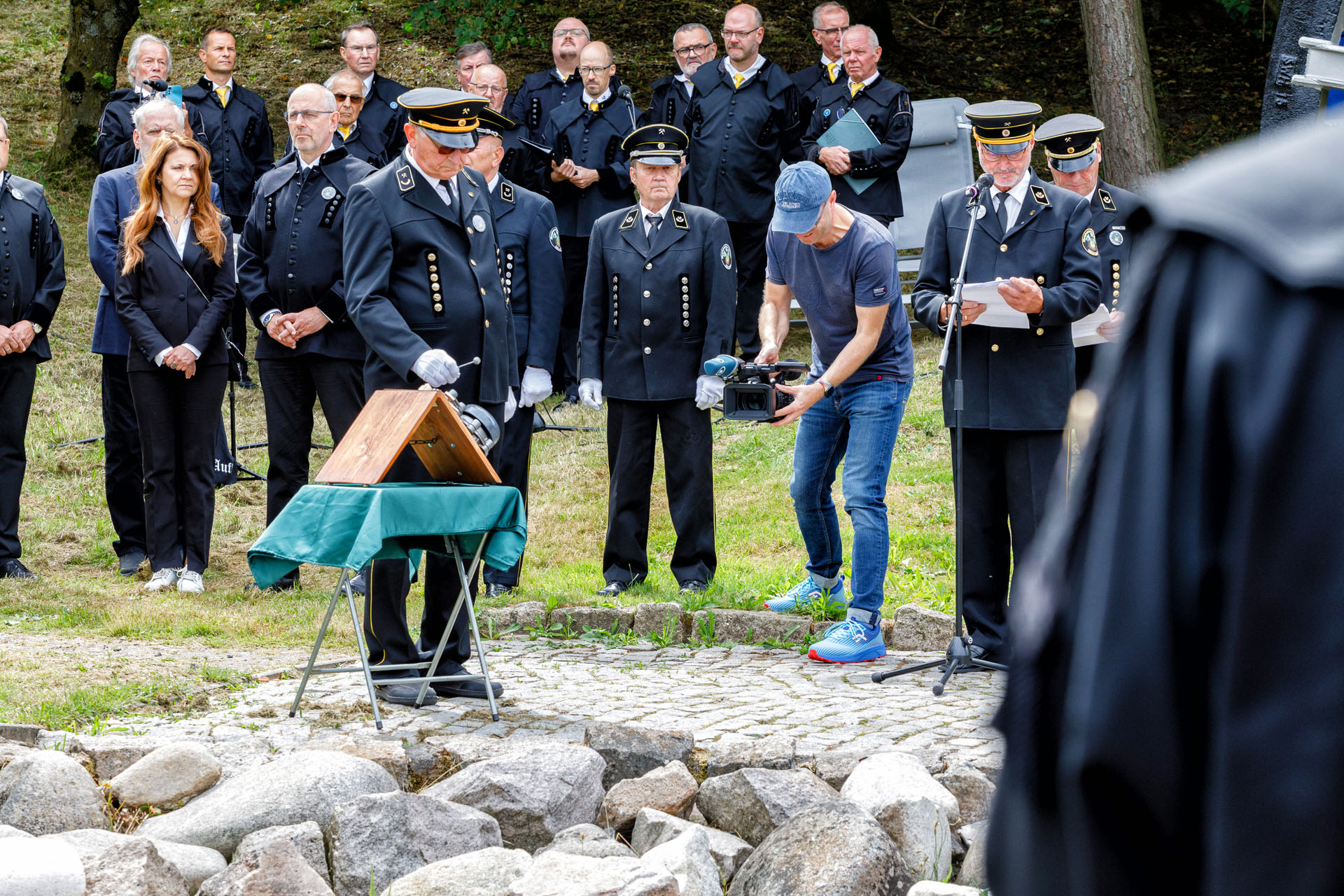 Ein Mann im Habit schlägt eine Signalglocke, ein Mann liest am Mikrofon von einem Zettel ab
